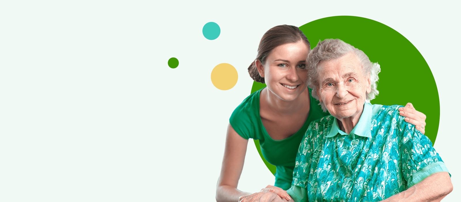 Ältere Frau mit gemustertem türkisfarbenem Hemd sitzt, eine jüngere Frau in grünem T-Shirt steht hinter ihm und legt die Hand auf seine Schulter.