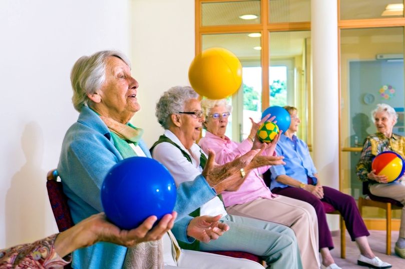 Mehrere ältere Menschen sitzen in einem Raum und halten bunte Bälle in den Händen, einige werfen sie in die Luft.